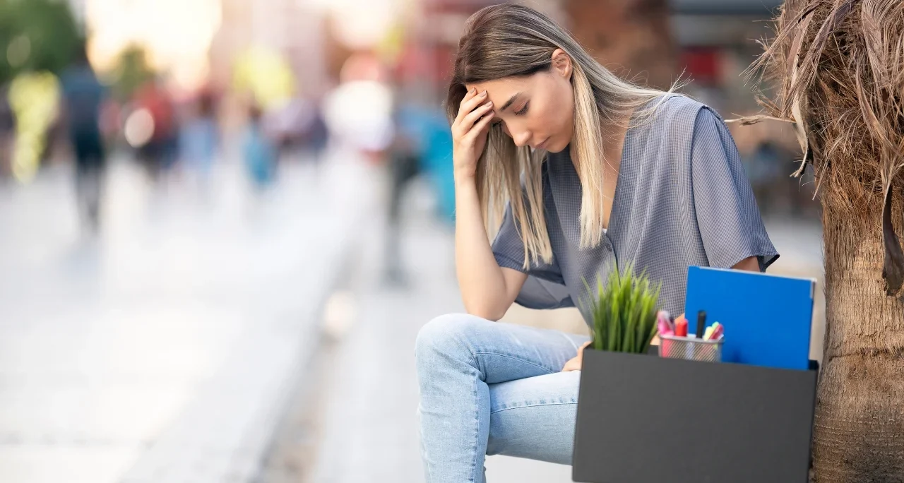 Junge Frau sitzt draußen, sieht nachdenklich aus und hält ihren Kopf, neben ihr eine Box mit Büromaterialien. Symbolisiert Arbeitslosigkeit und neue Chancen durch Weiterbildung.