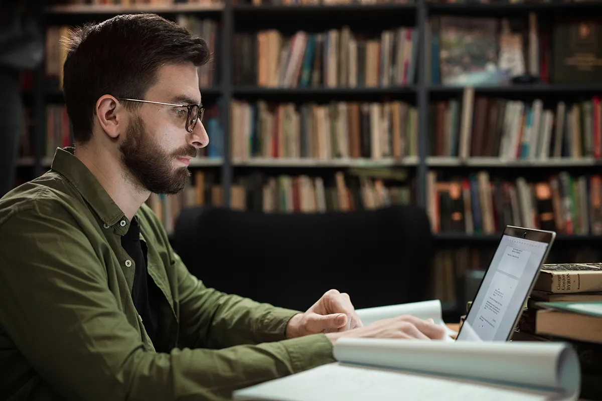 Mann mit Brille und grünem Hemd arbeitet am Laptop in einer Bibliothek, möglicherweise auf der Suche nach Informationen für BOS-Abbrecher.